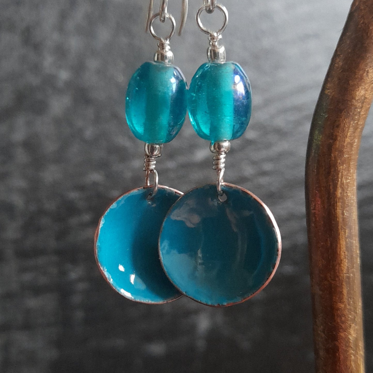 Cloe up of a pair of handmade blue enamel bowl dangle earrings, presented with a blue oval glass bead, hanging from a copper hoop, set on silver earwires.