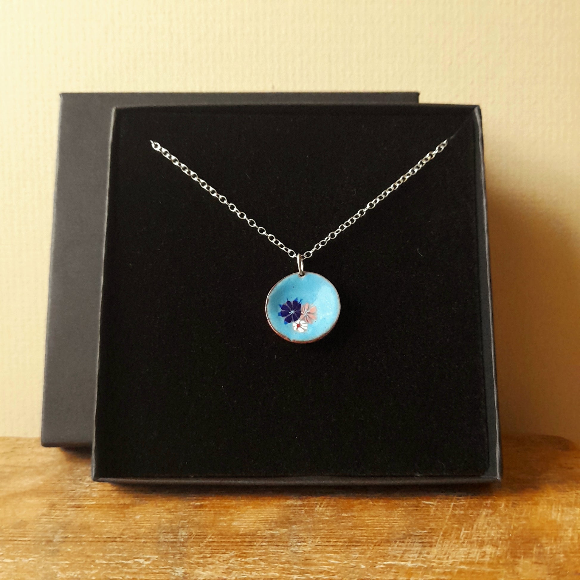 Pale blue circular enamel pendant, with 3 small pretty flowers in blue, white and pink left of centre . the necklace is silver chain and it is presented in a black gift box.