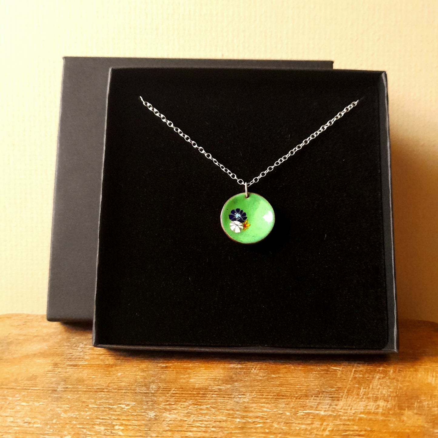 Grass green circular enamel pendant, with 3 small pretty flowers in blue, white and yellow left of centre . the necklace is silver chain and it is presented in a black gift box.