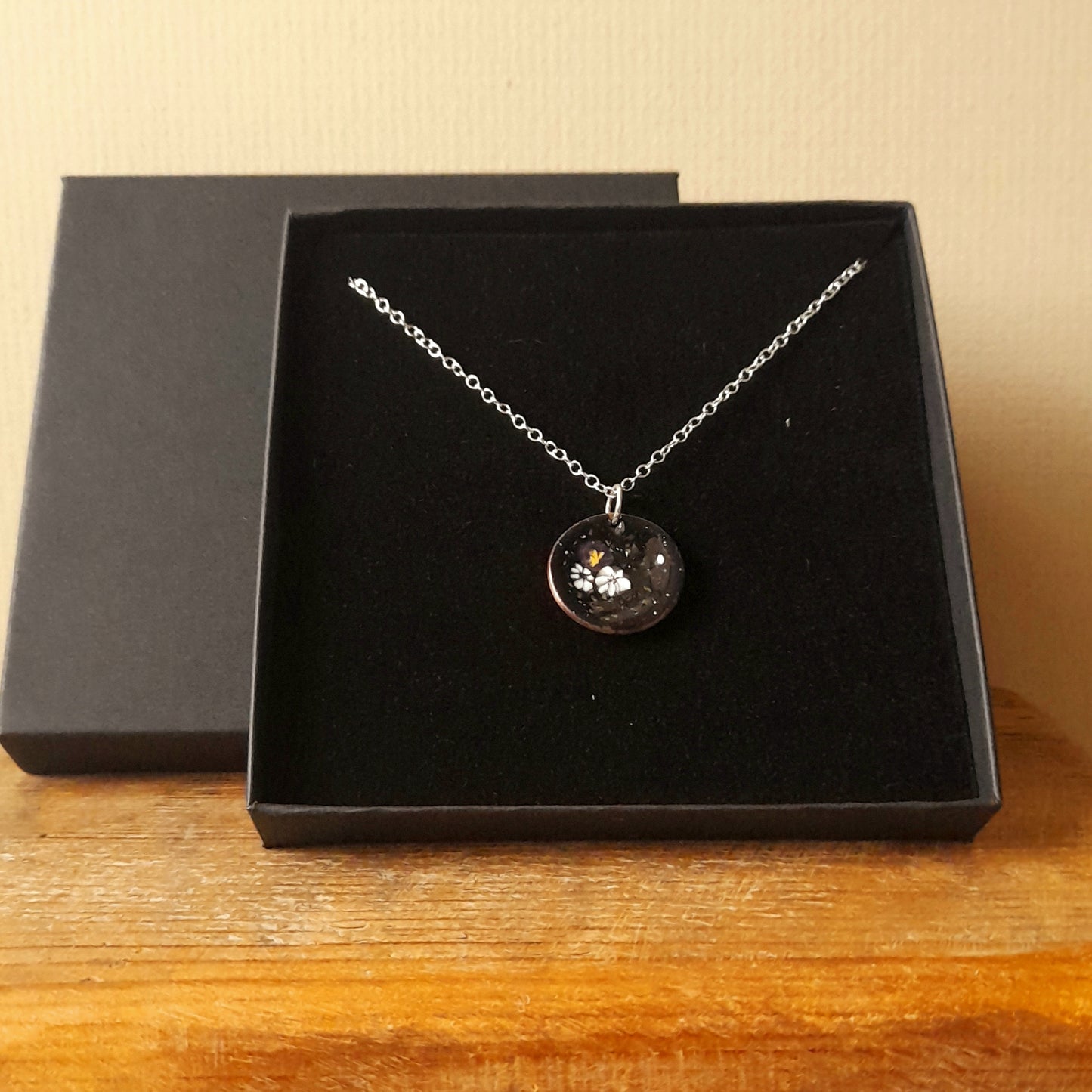 Black circular enamel pendant, with 3 small pretty flowers in white and red left of centre . The necklace is silver chain and it is presented in a black gift box.