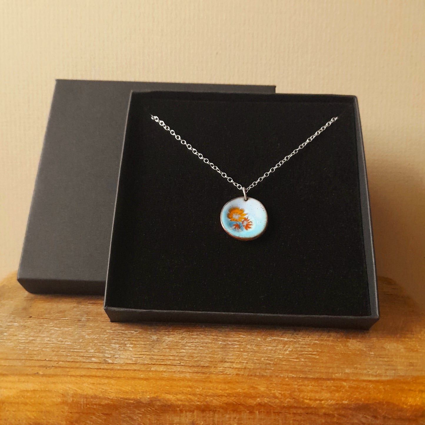 Very pale blue enamel pendant, with 3 small pretty flowers in orange, blue and red, left of centre . The necklace is silver chain and it is presented in a black gift box.