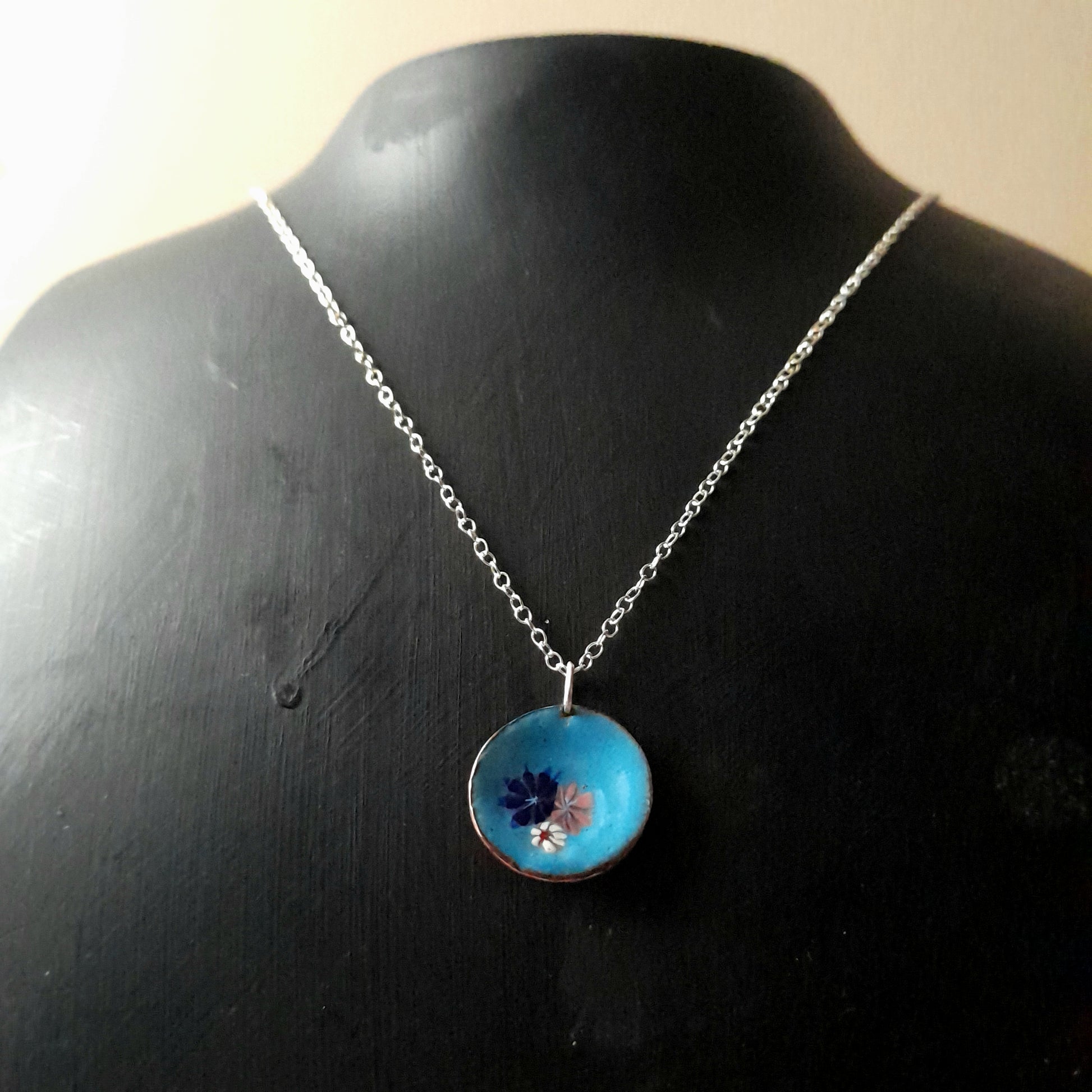 Mid blue enamel pendant, with 3 small pretty flowers in white, blue and pink, left of centre . The necklace is on silver chain is hown on a black background.