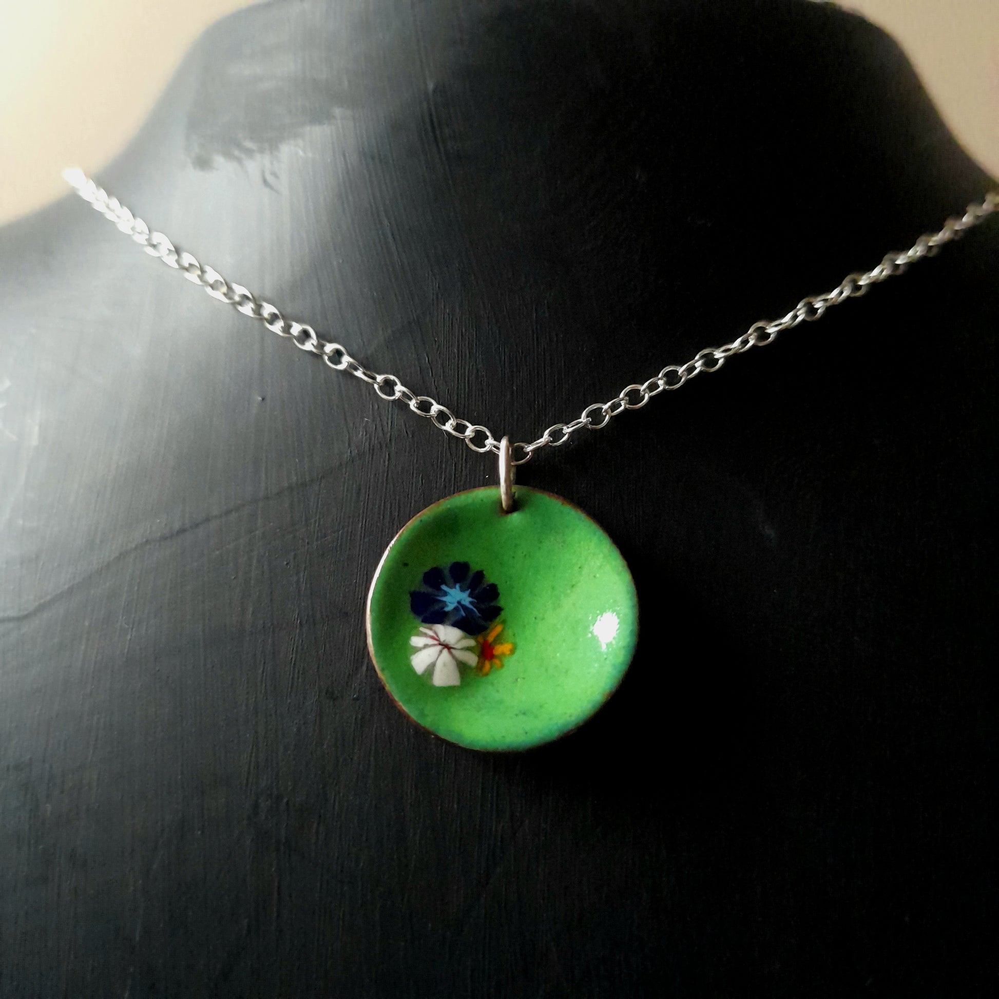 Grass green circular enamel pendant, with 3 small pretty flowers in blue, white and yellow left of centre . the necklace is silver chain and it is presented in a black gift box.