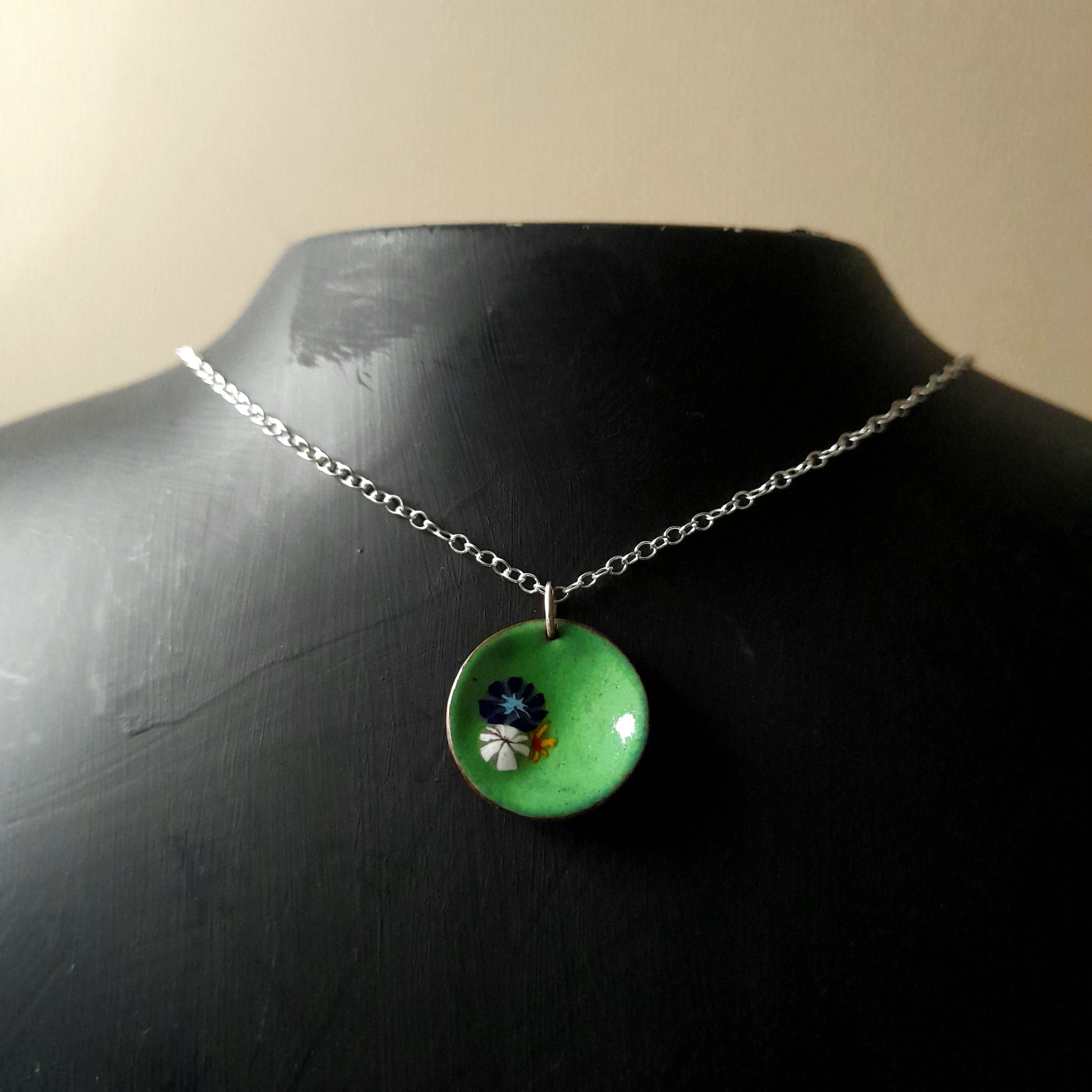 Grass green circular enamel pendant, with 3 small pretty flowers in blue, white and yellow left of centre . the necklace is on silver chain and it is presented on a black background.