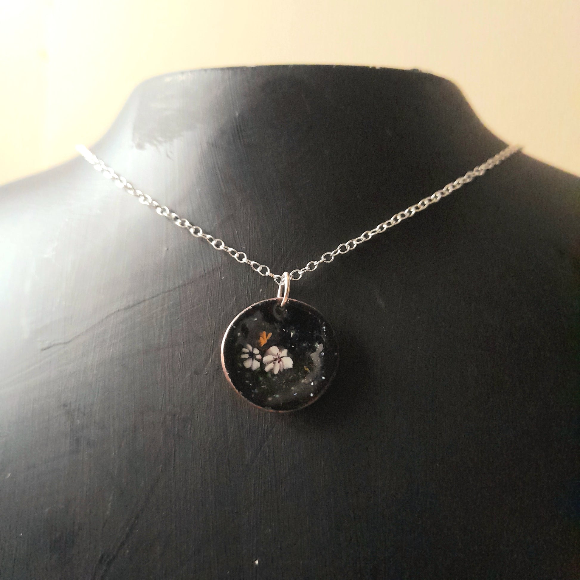 Black circular enamel pendant, with 3 small pretty flowers in white and yellow, left of centre . The necklace is on silver chain and it is presented on a black background.