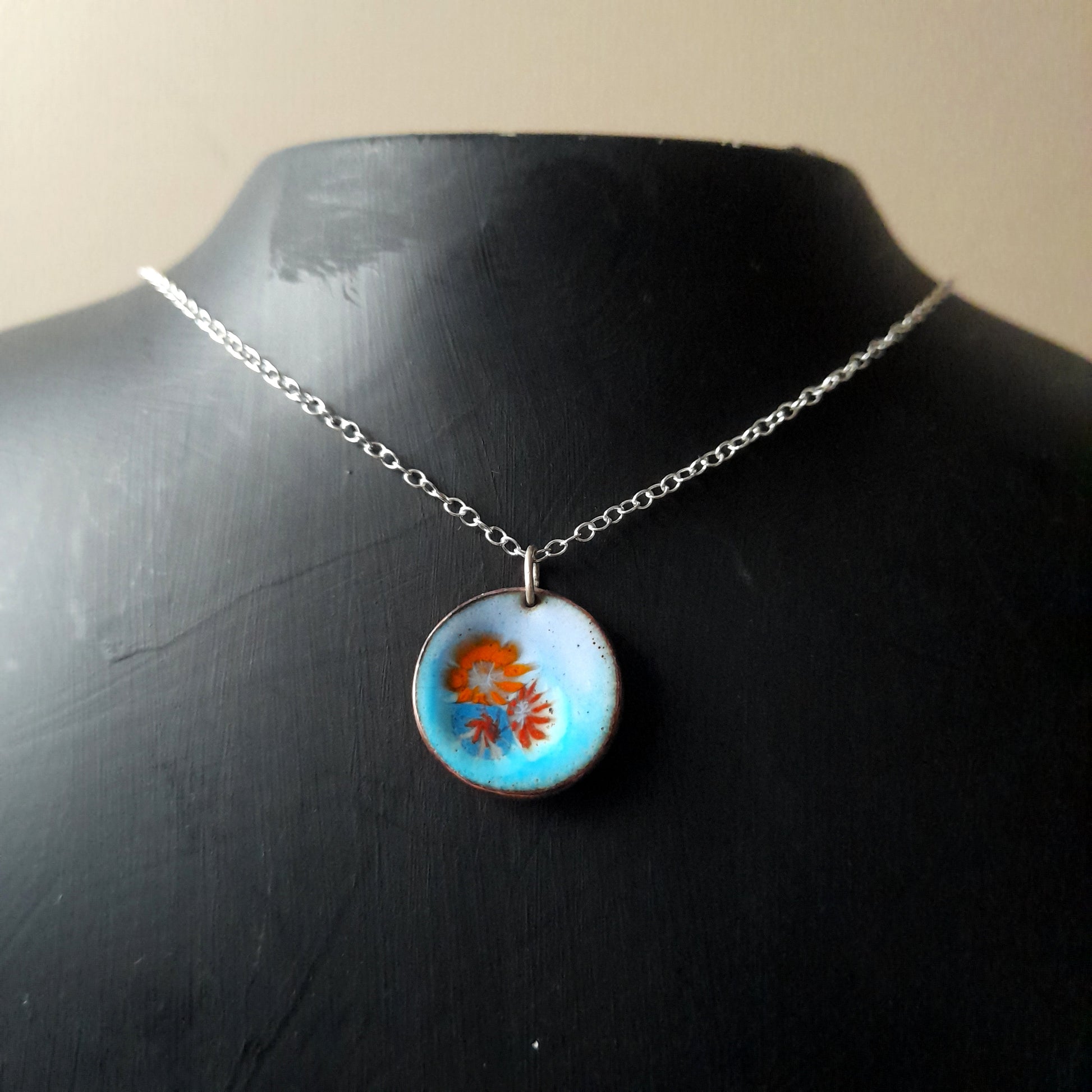 Very pale blue enamel pendant, with 3 small pretty flowers in orange, blue and red, left of centre . The necklace is on silver chain and it is presented on a black background.