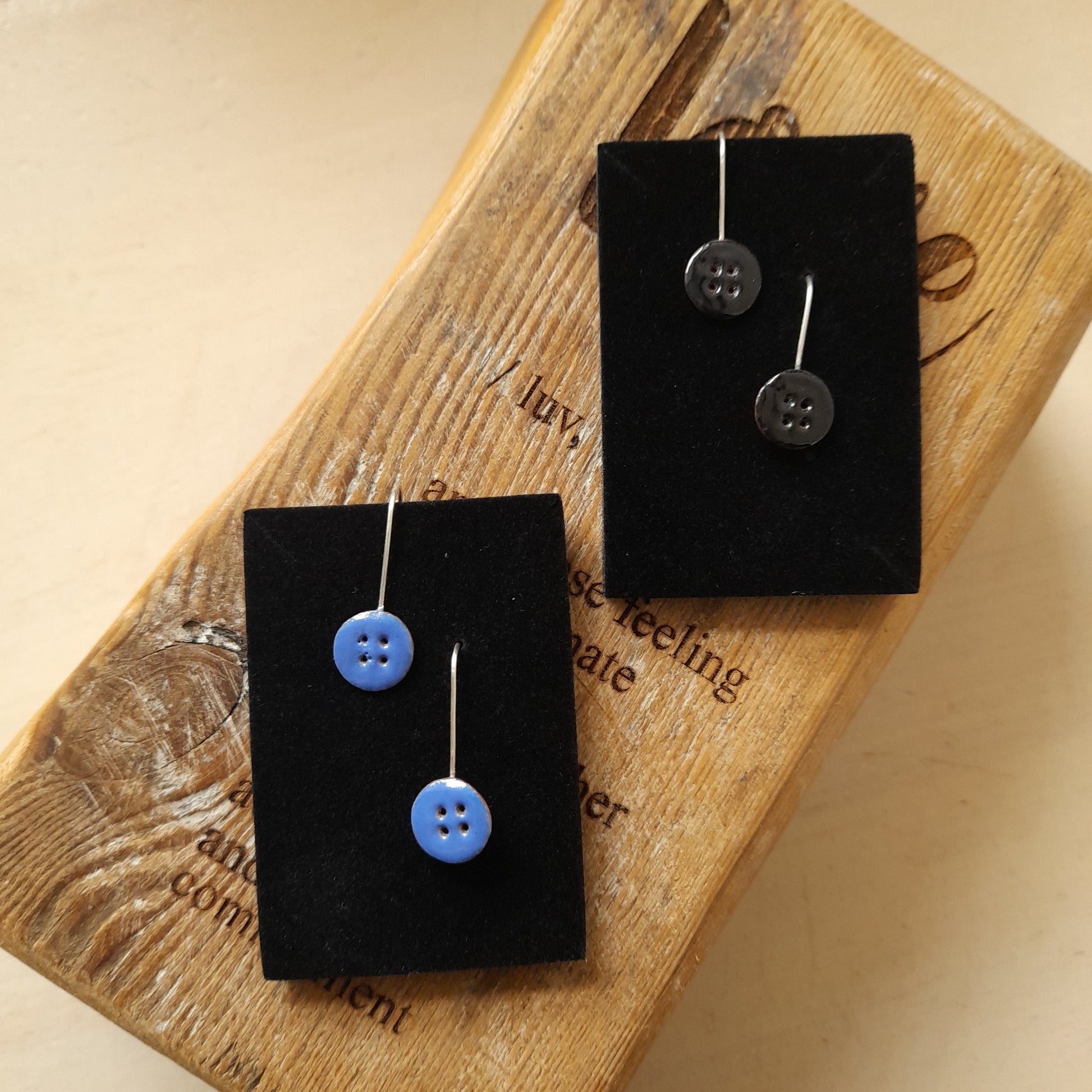 2 pairs of handmade button drop earrings, in blue and black with 4 holes on each. Displayed on a wood background.