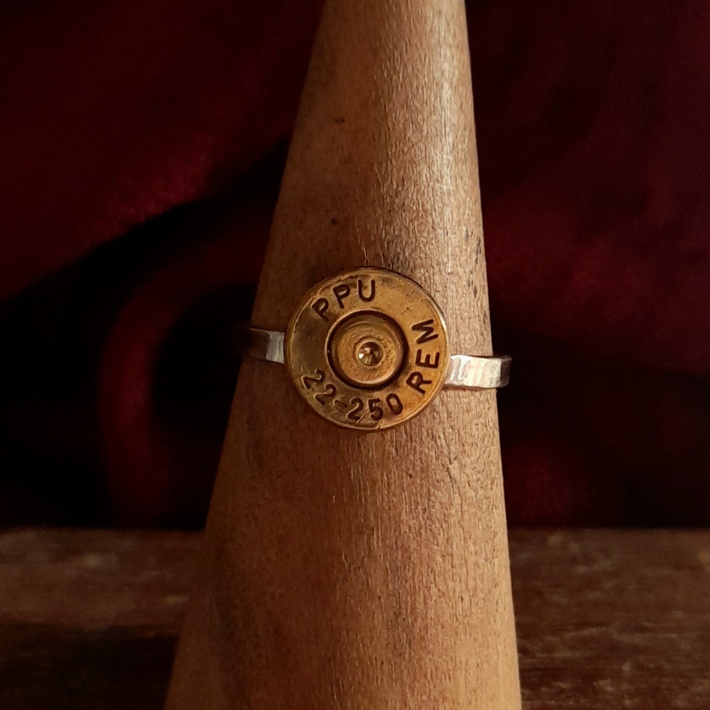 This image shows 1 cartridge ring in close up. It has a silver band with the bottom of the cartridge soldered on like a signet ring, there is writing on the cartridge ends. They are on a grainy wood background.