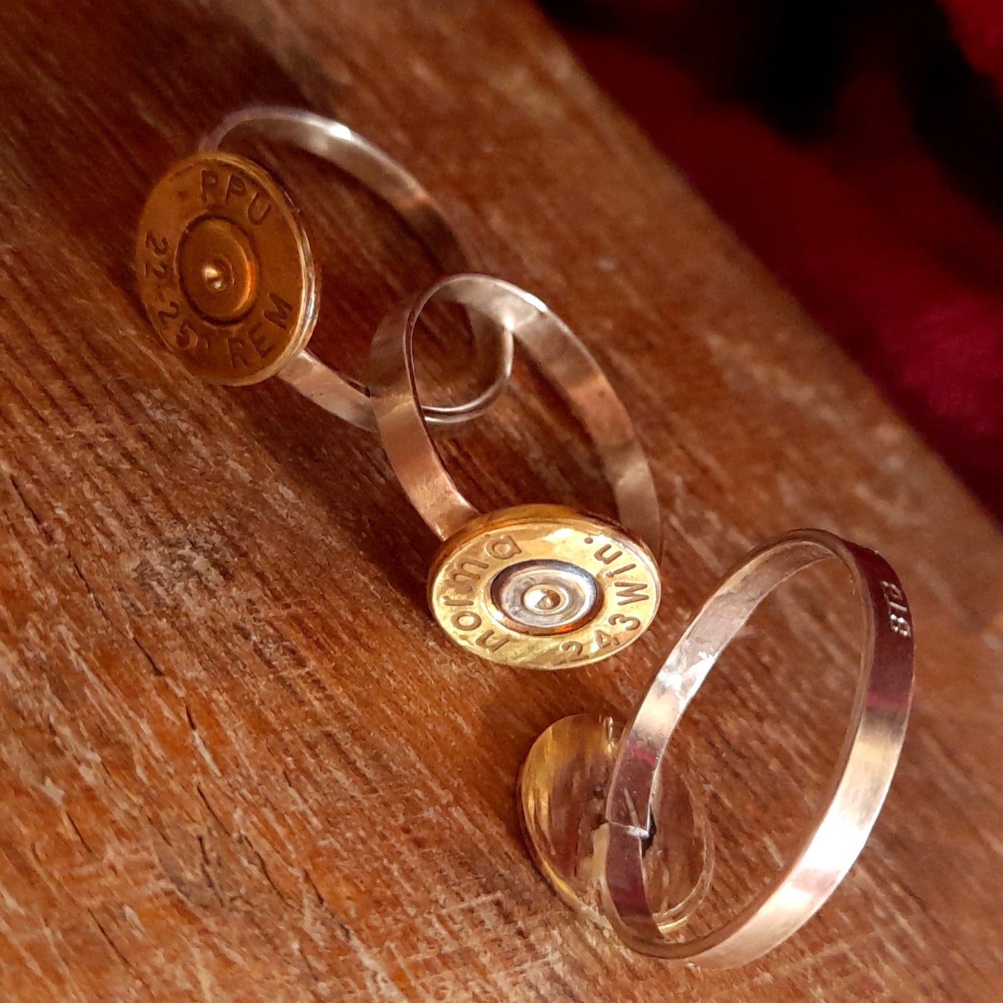 This image shows 3 cartridge rings in close up. They have copper bands with the bottom of the cartridge soldered on like a signet ring, there is writing on the cartridge ends. they are on a grainy wood background