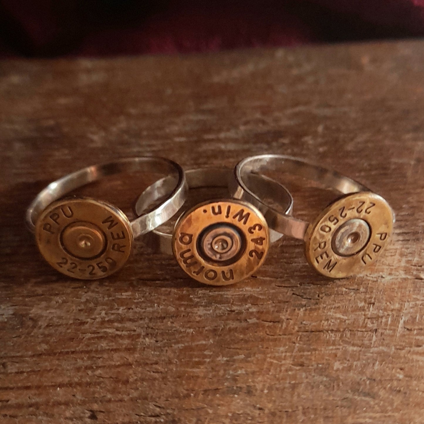 This image shows 3 cartridge rings in close up. They have silver bands with the bottom of the cartridge soldered on like a signet ring, there is writing on the cartridge ends. they are on a grainy wood background