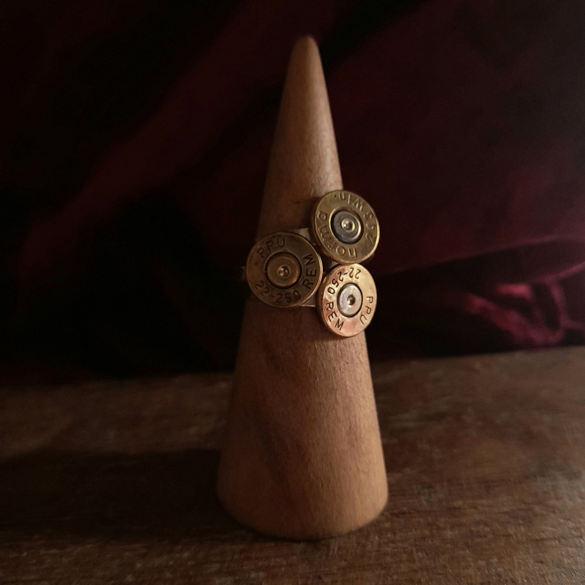 This image shows 3 cartridge ring in close up. They have silver bands with the bottom of the cartridge soldered on like a signet ring, there is writing on the cartridge ends. They are on a grainy wood background.