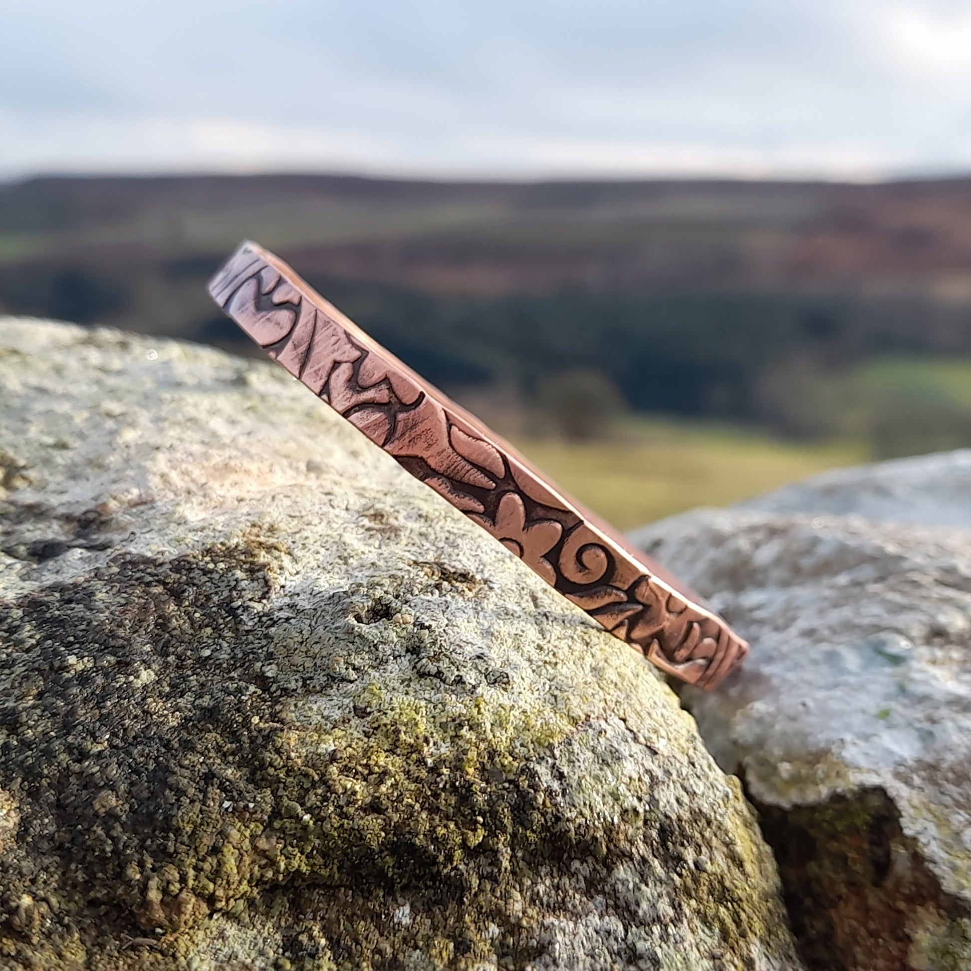 Floral embossed solid copper bangle, on white stone with countryside in the background 