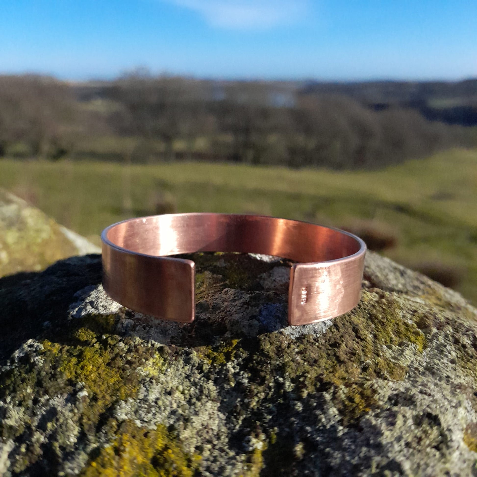 Plain wide, adjustable copper bangle in close up, set on a white rock, in the background is a countryside view