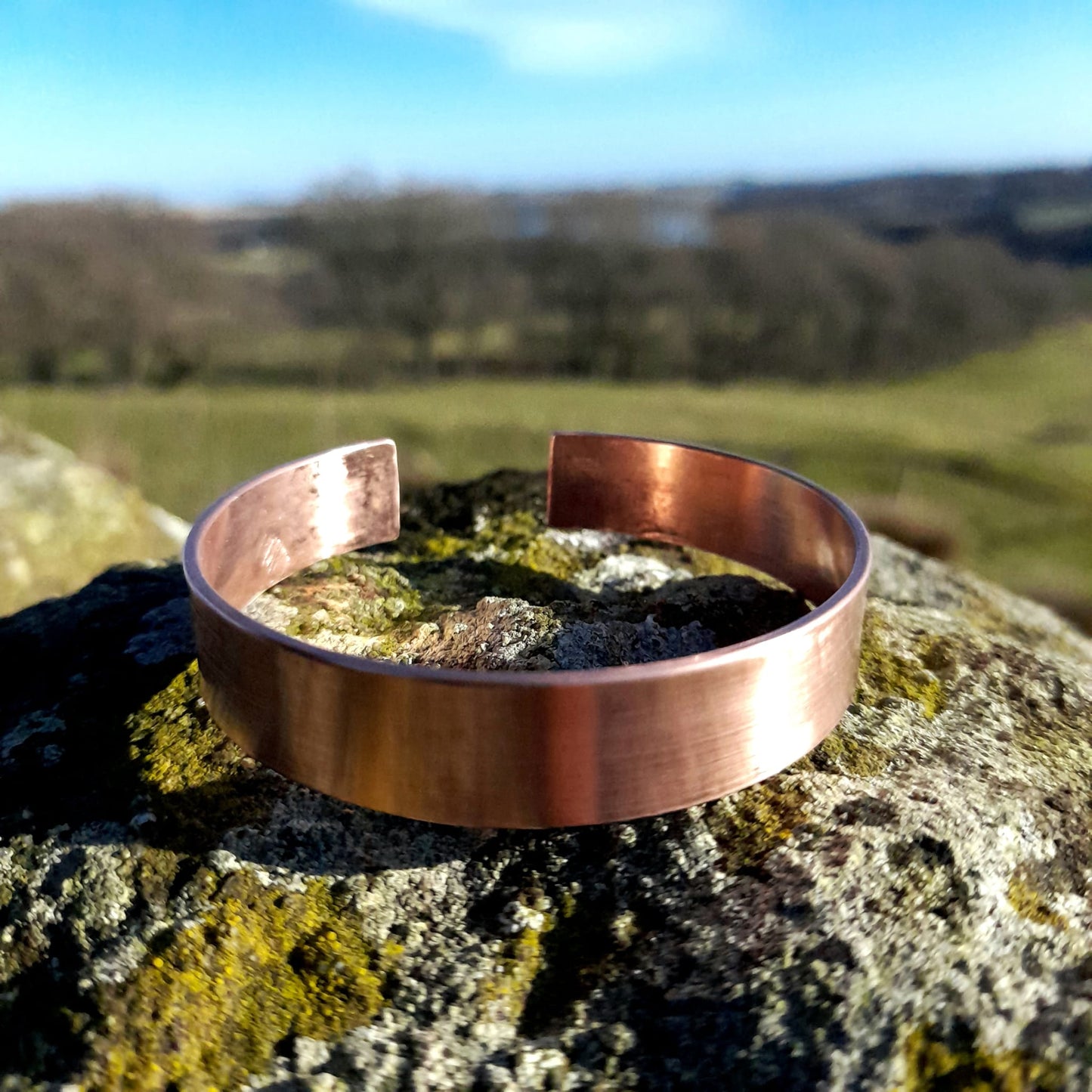 Plain wide, adjustable copper bangle in close up, set on a white rock, in the background is a countryside view