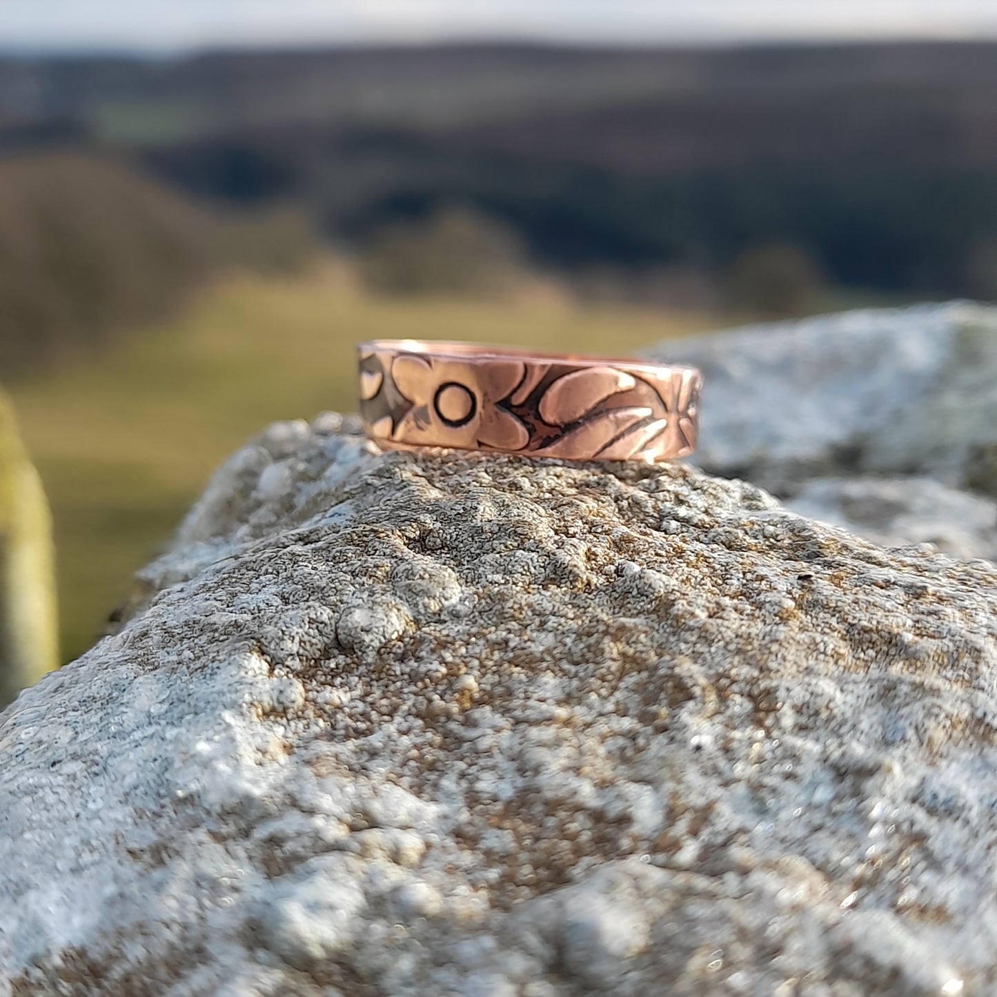 Chunky copper ring with floral patterned embossed on surface. Pictured on white rock with countryside in the background