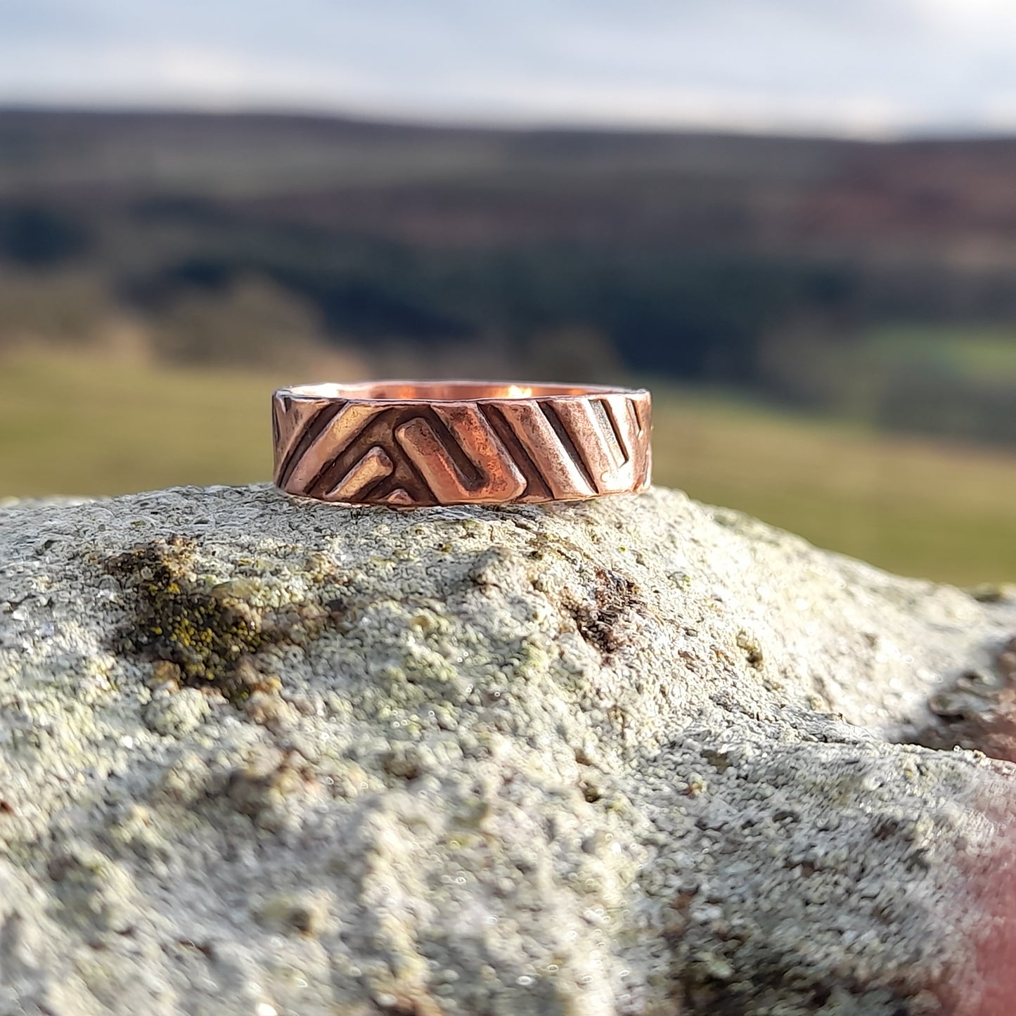 Chunky copper ring with geometric patterned embossed on surface. Pictured on white rock with countryside in the background