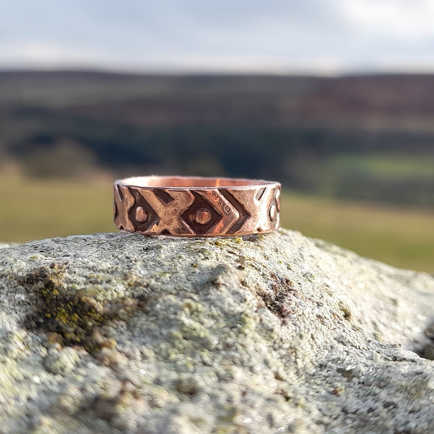 Chunky copper ring with geometric patterned embossed on surface. Pictured on white rock with countryside in the background