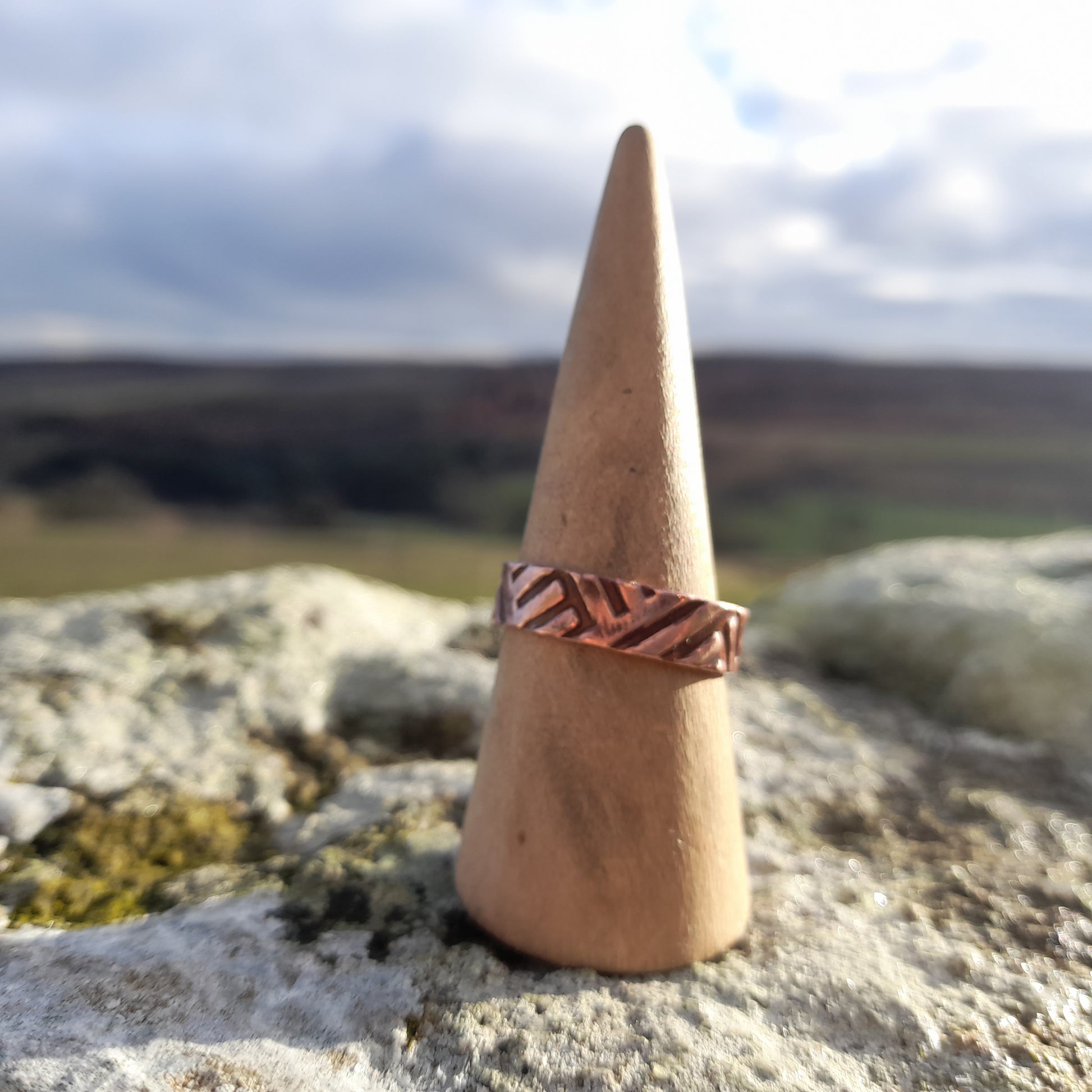 Chunky copper ring with geometric patterned embossed on surface. Pictured on ring cone on a white rock with countryside in the background