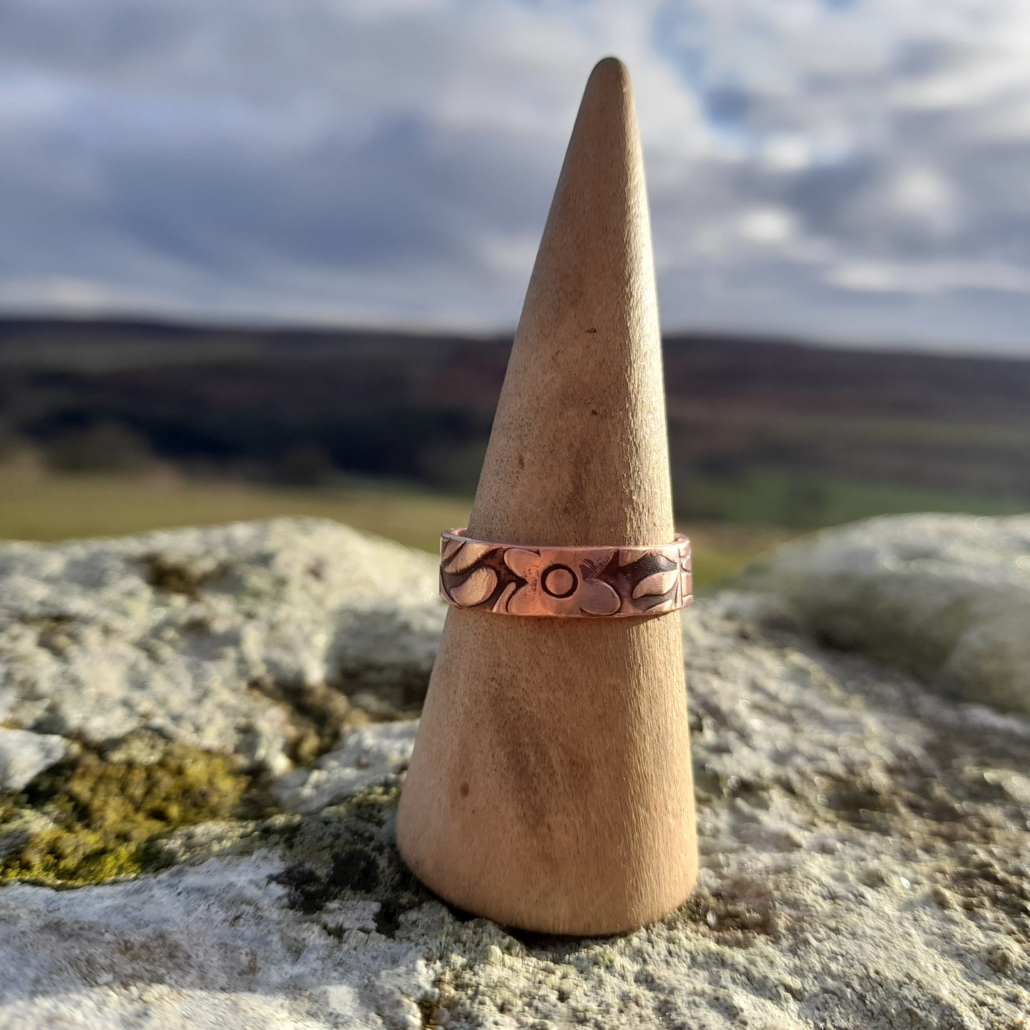 Chunky copper ring with floral patterned embossed on surface. Pictured on ring cone on a white rock with countryside in the background