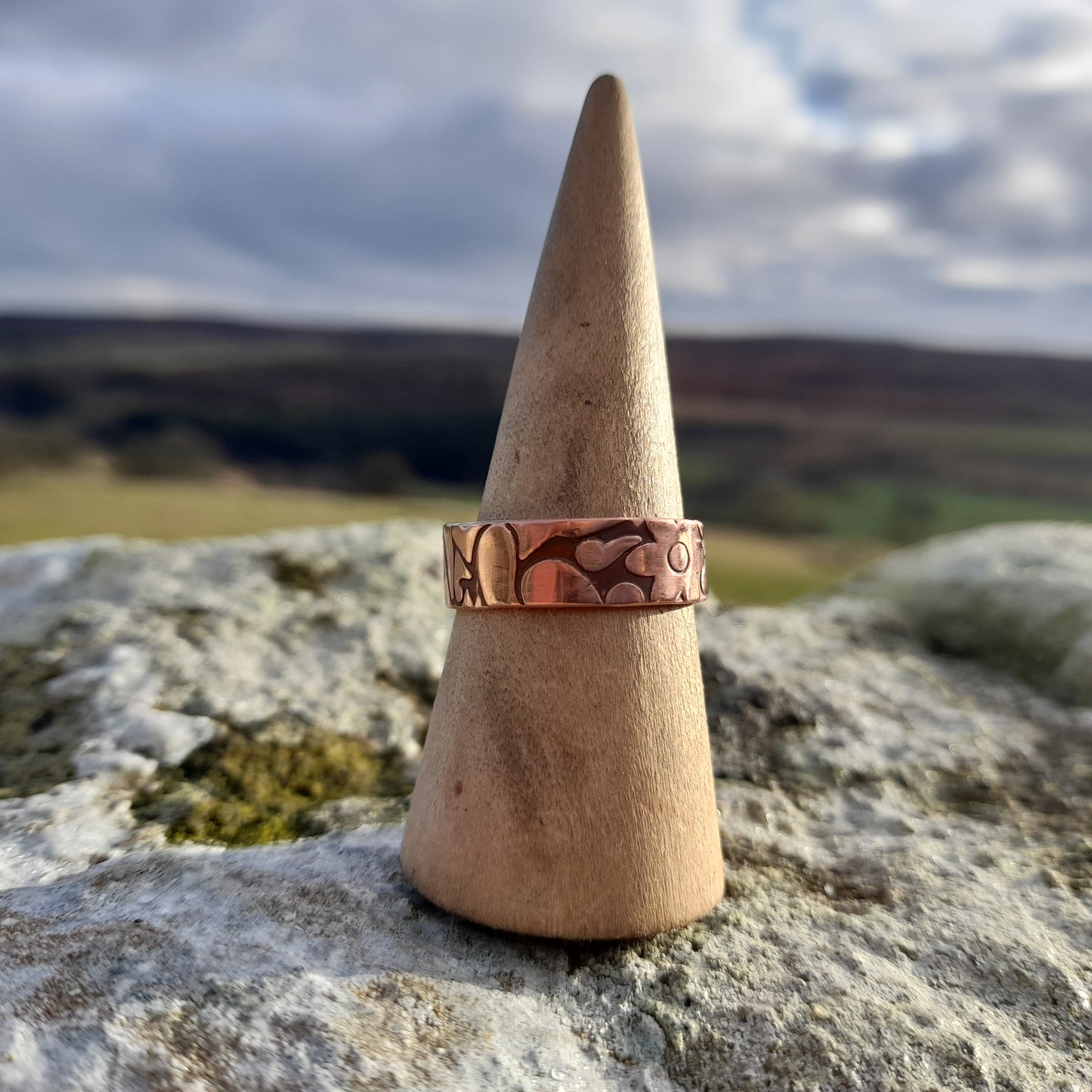 Chunky copper ring with geometric patterned embossed on surface. Pictured on ring cone on a white rock with countryside in the background