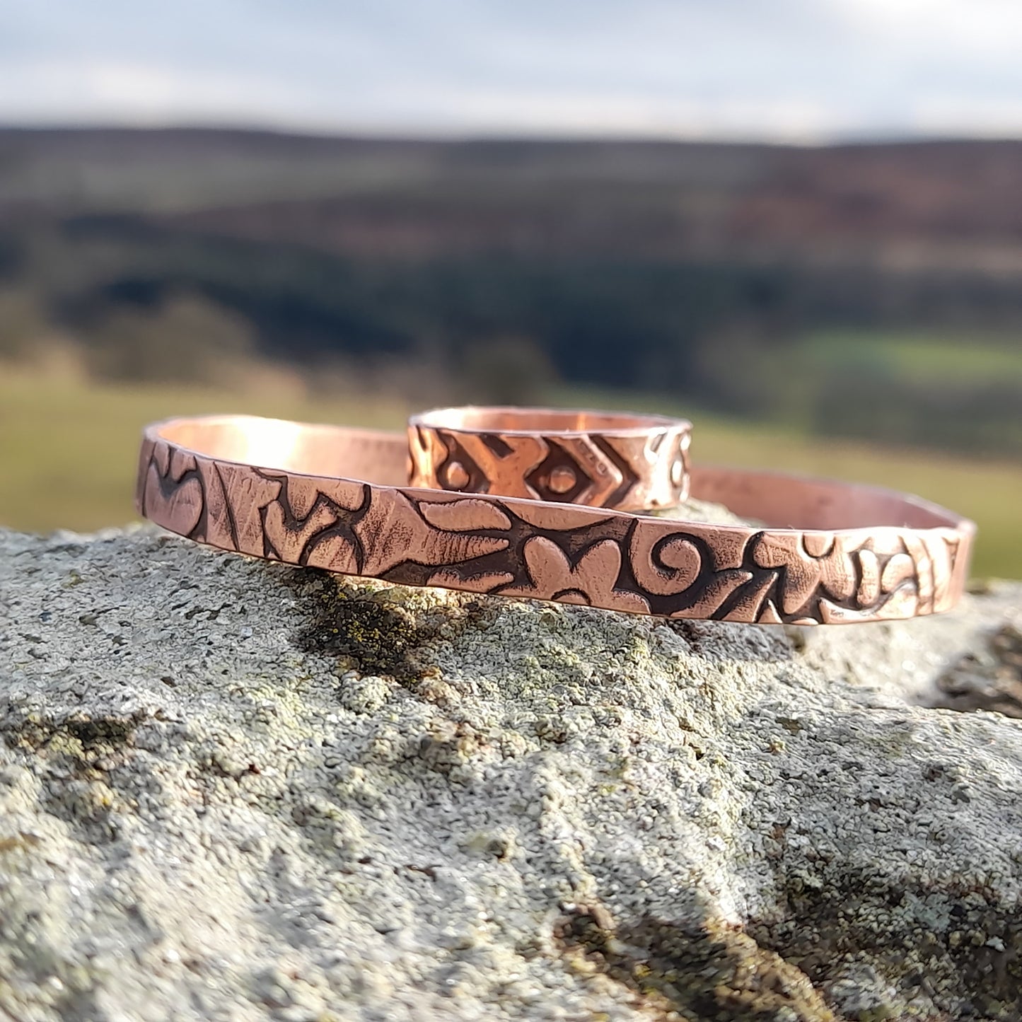 Chunky copper ring with geometric pattenr and bangle with floral embossed design on surface. Pictured on white rock with countryside in the background