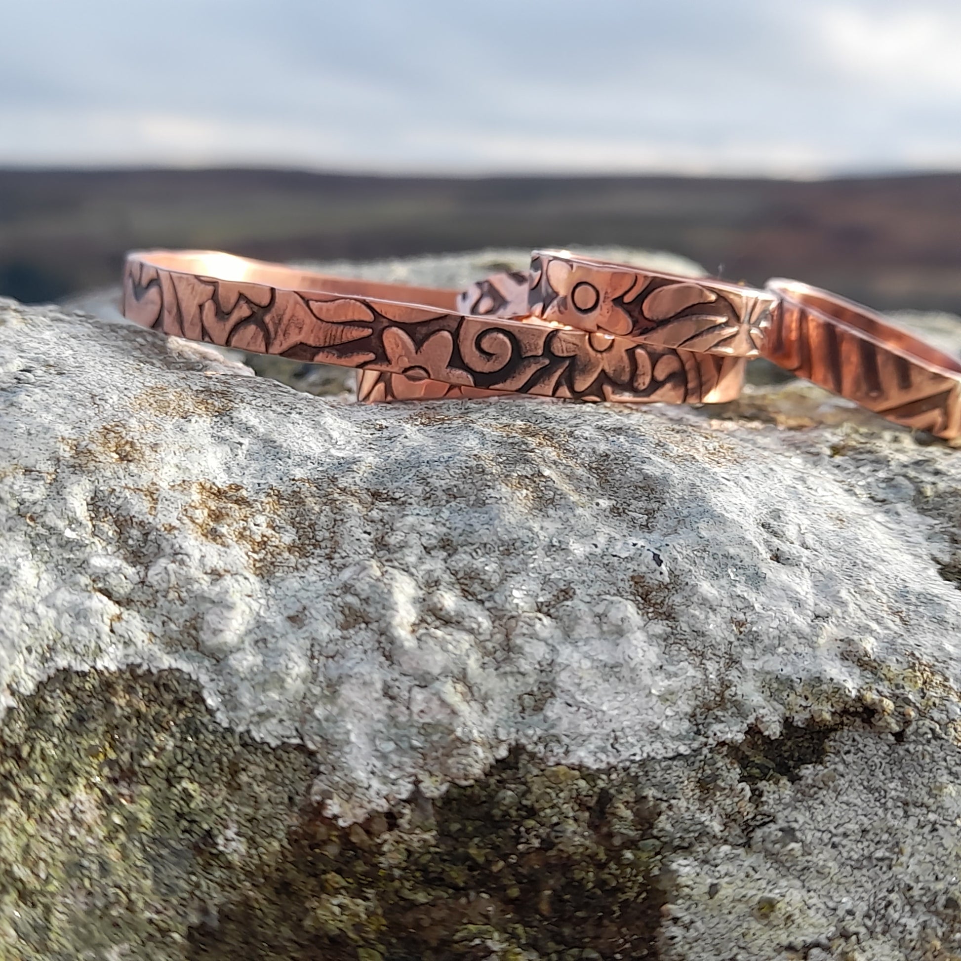 Floral embossed solid copper bangles and rings on white stone with countryside in the background 