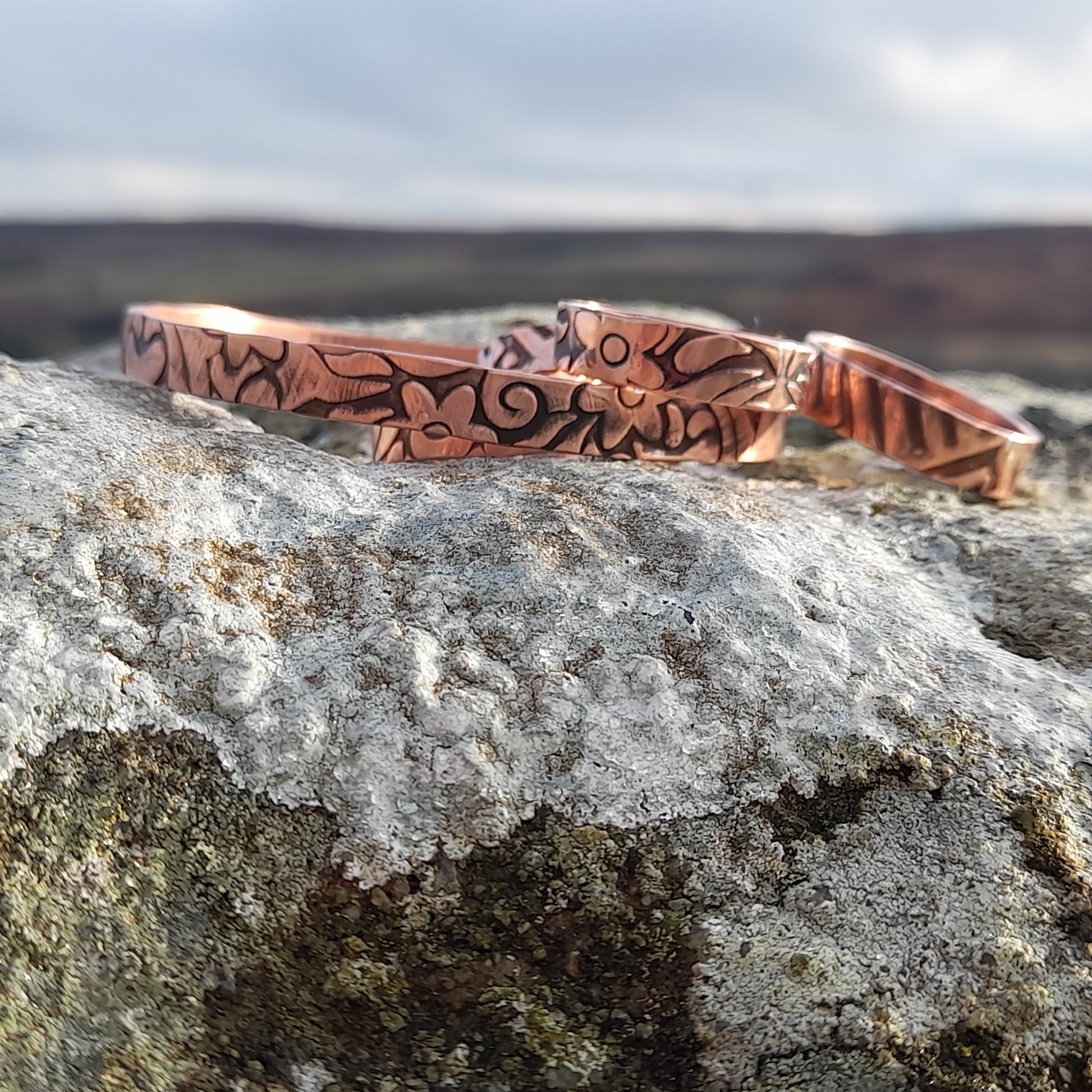 Floral embossed solid copper rings and bangle, on white stone with countryside in the background 