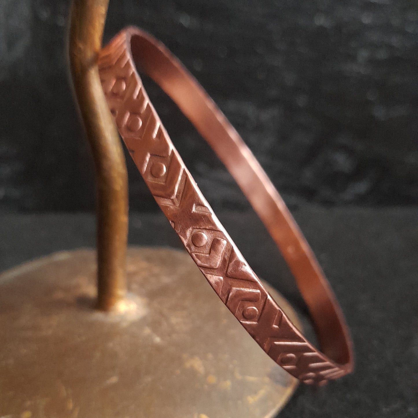 Copper round bangle leaning on a candlestick, showing the geometric embossed design