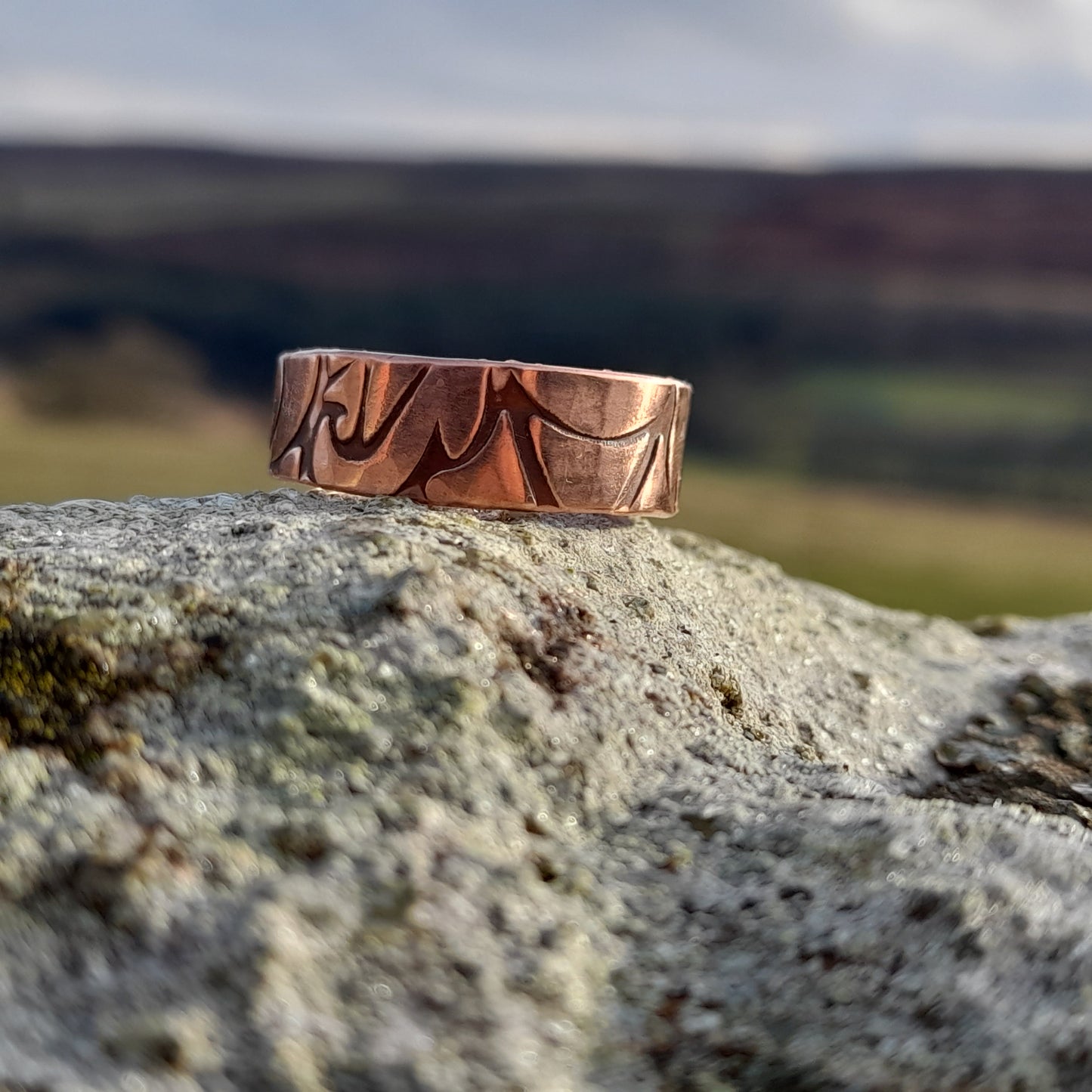 Chunky copper ring with floral patterned embossed on surface. Pictured on white rock with countryside in the background