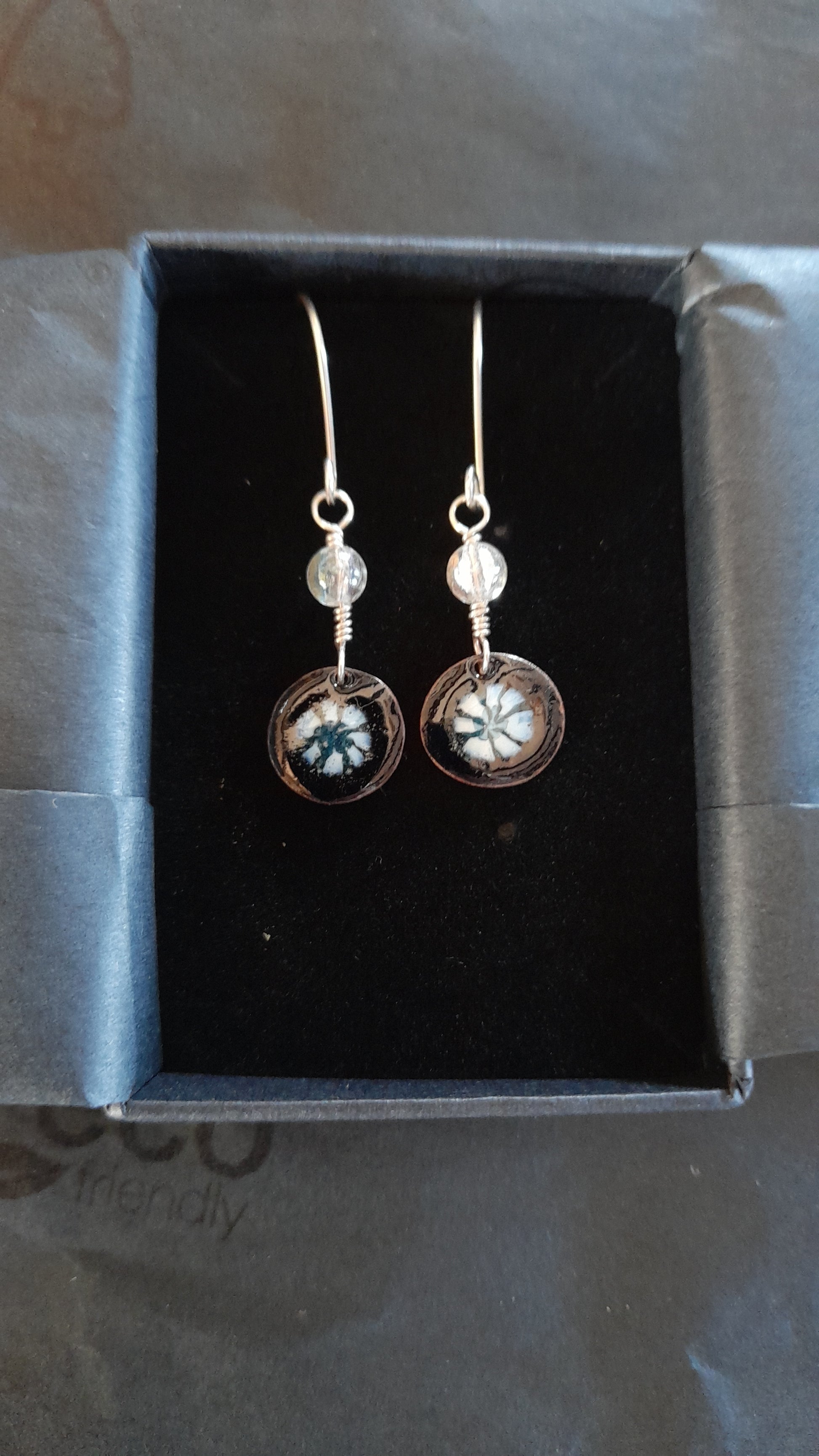 This photo shows a pair of black dangle enamel earrings with a pretty white and blue floral design in the centre, they are on silver earwires. The items are packed in a black gift box with tissue paper.