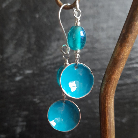 A pair of handmade blue enamel bowl dangle earrings, presented with blue oval glass bead, hanging from a copper hoop, set on silver earwires.