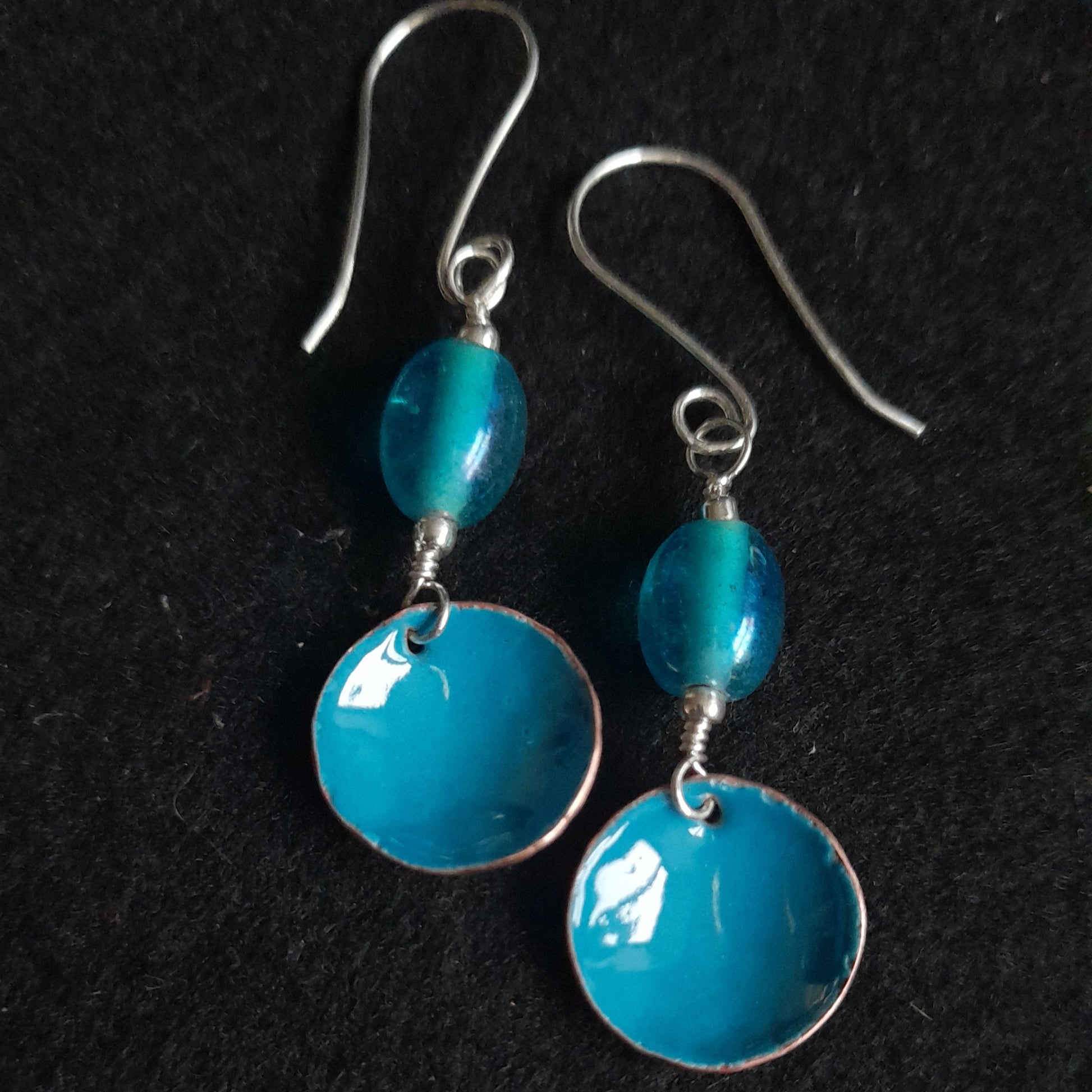 A pair of handmade blue enamel bowl dangle earrings, presented with a blue oval glass bead, hanging from a copper hoop, set on silver earwires.