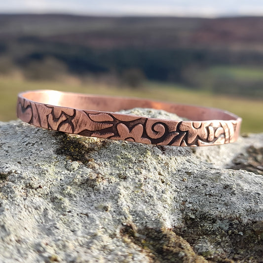 Floral embossed solid copper bangle, on white stone with countryside in the background 