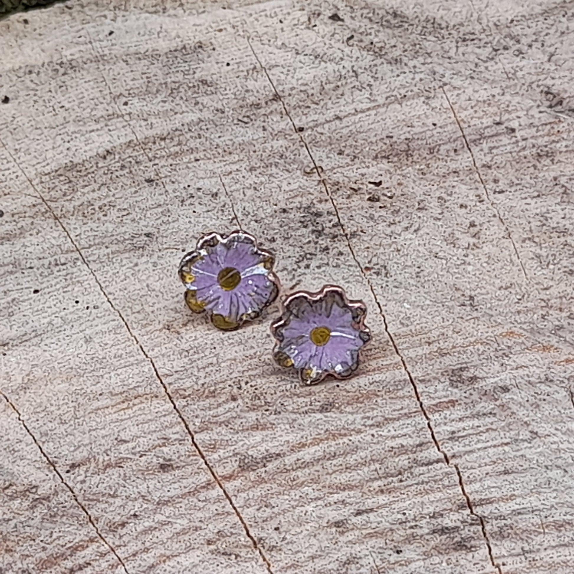 Light purple, mauve flower shaped stud earrings. The edges are fluted creating 8 petal shapes. The centre is yellow and there is hand painted detail to the petals. 